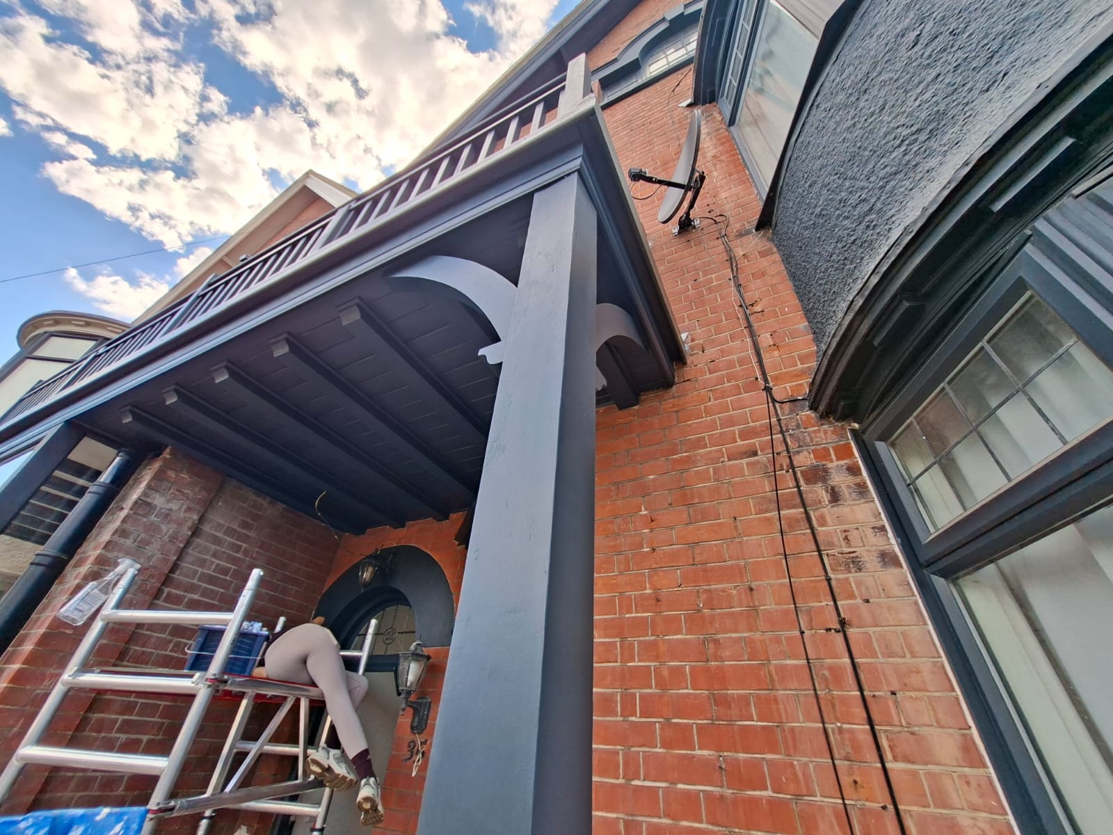 Looking up at the porch canopy and balcony soffit of a red-brick period house mid-repaint